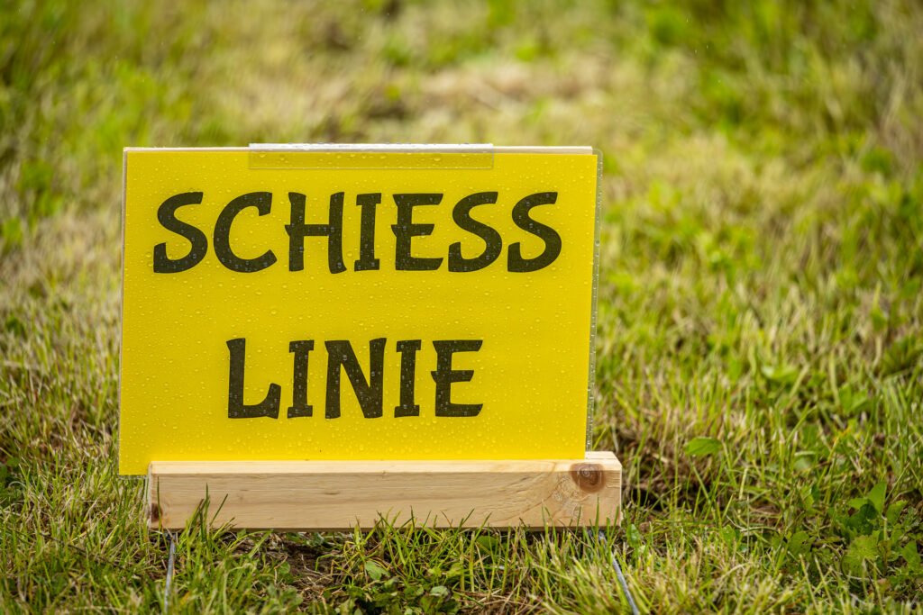Gelbes Schild mit der Aufschrift Schiesslinie am Rand der Wiese